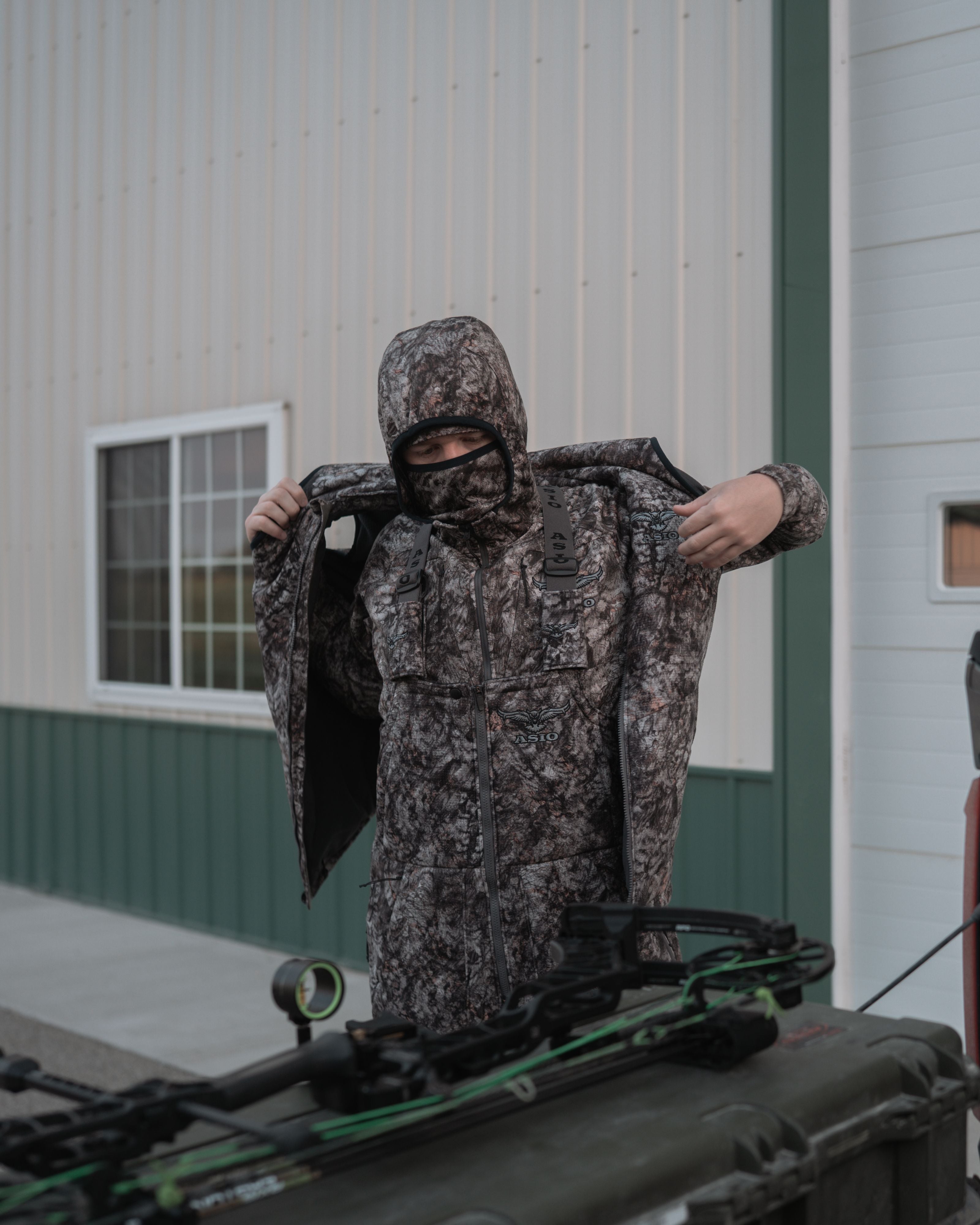 Hunter pulling on the ASIO Gear heavyweight vest in Raptor camo over cold-weather gear next to a truck bed.