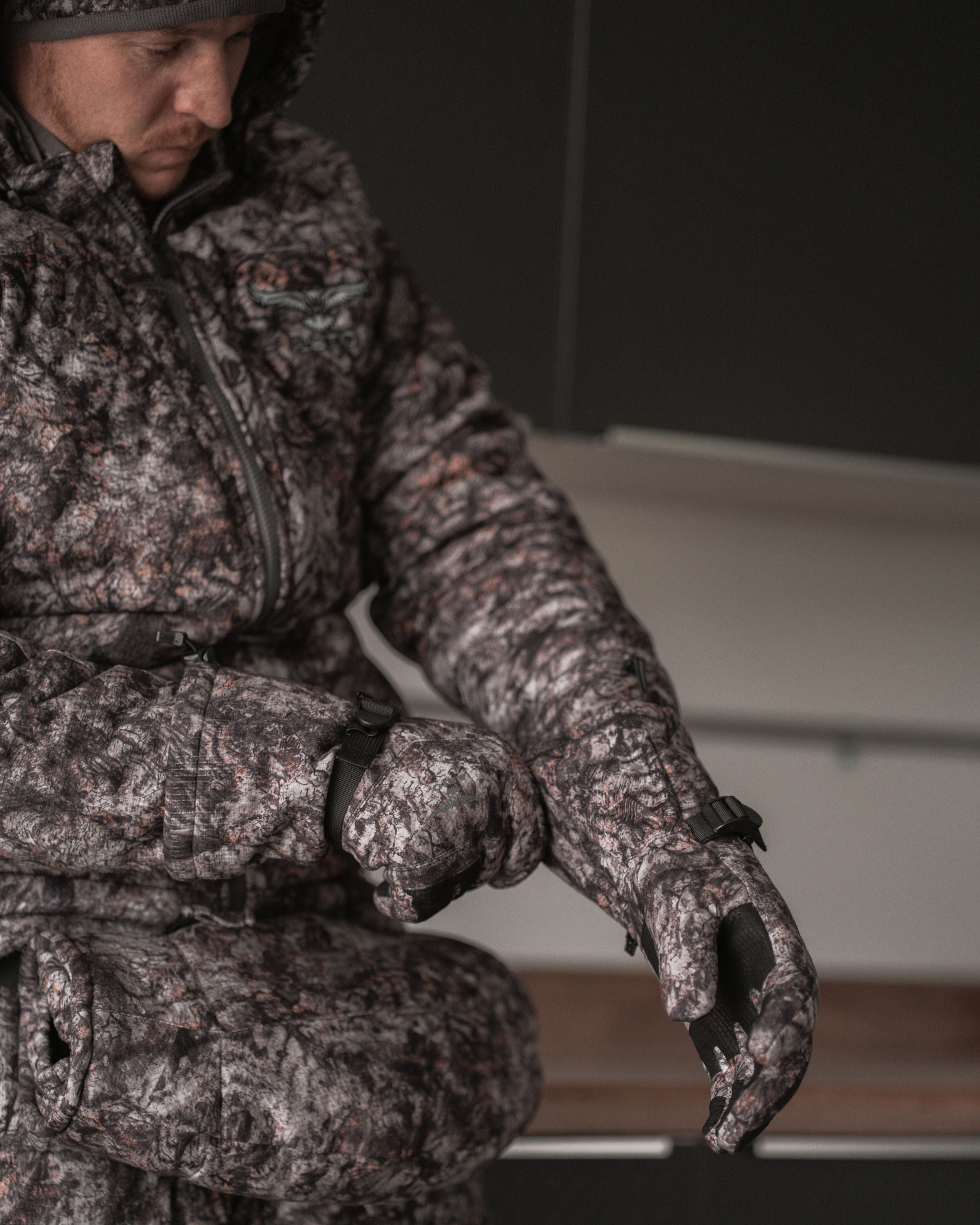 Hunter wearing ASIO Gear RAPTOR camo heavyweight insulated gloves securing wrist fit indoors before cold-weather bowhunt