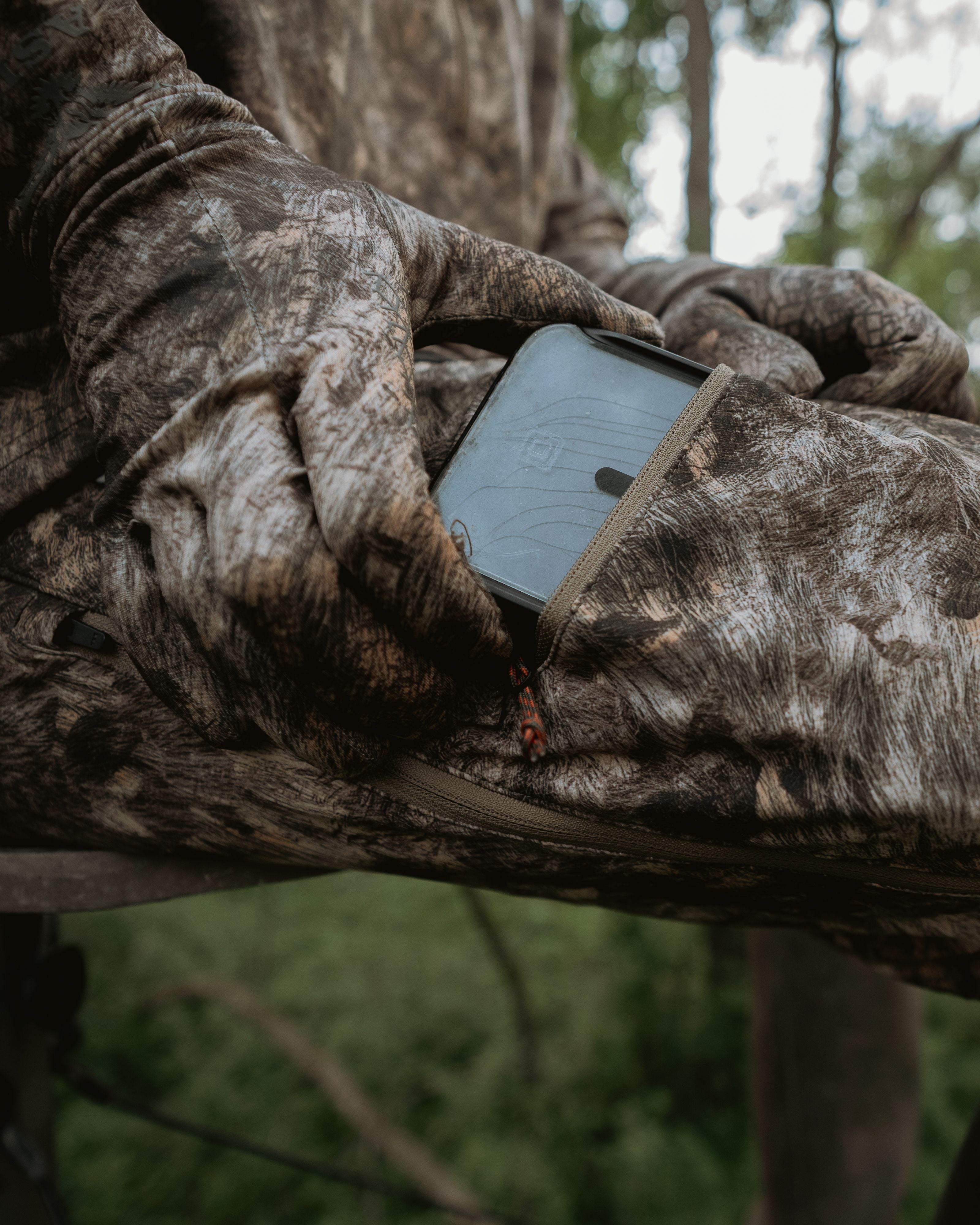 Hunter accessing phone from zippered thigh pocket on ASIO Gear lightweight pant in RAPX camo while seated in treestand