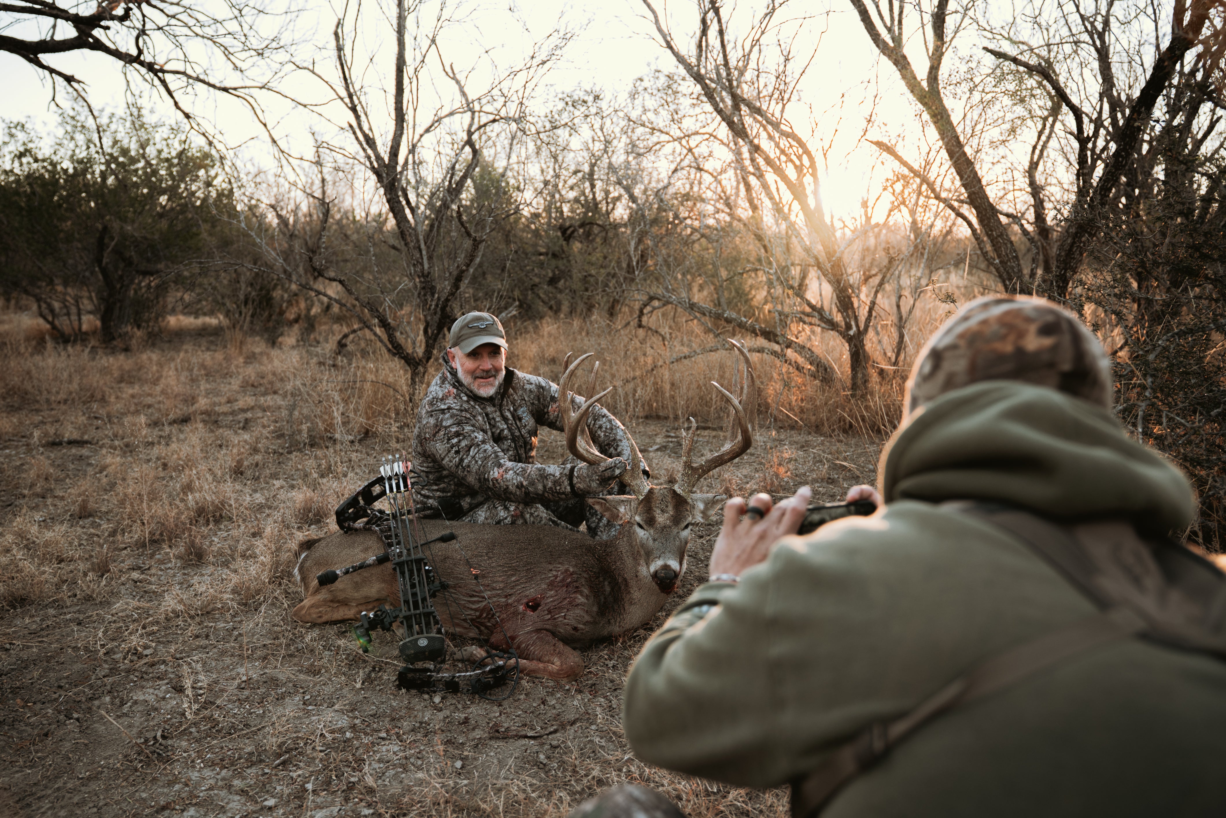 ASIO Gear bowhunter in RAPTOR camo poses with a mature whitetail buck at sunrise, while a hunting partner captures the moment in the Texas brush.